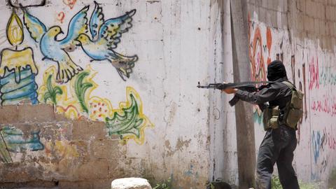 Ein Mitglied der Al-Qassam Brigade zielt mit einem Gewehr an einer Mauer in Gaza Stadt. Auf der Mauer ist ein Graffiti mit bunten Tauben als Symbol für Frieden zu sehen.