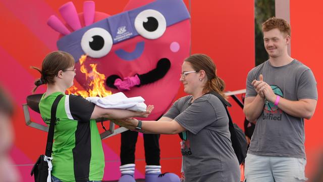 Eine Frau übergibt einer anderen Frau die gefaltete Fahne der Special Olympics. Im Hintergrund stehen das herzförmige Maskottchen der Special Olympics und ein Mann der klatscht.