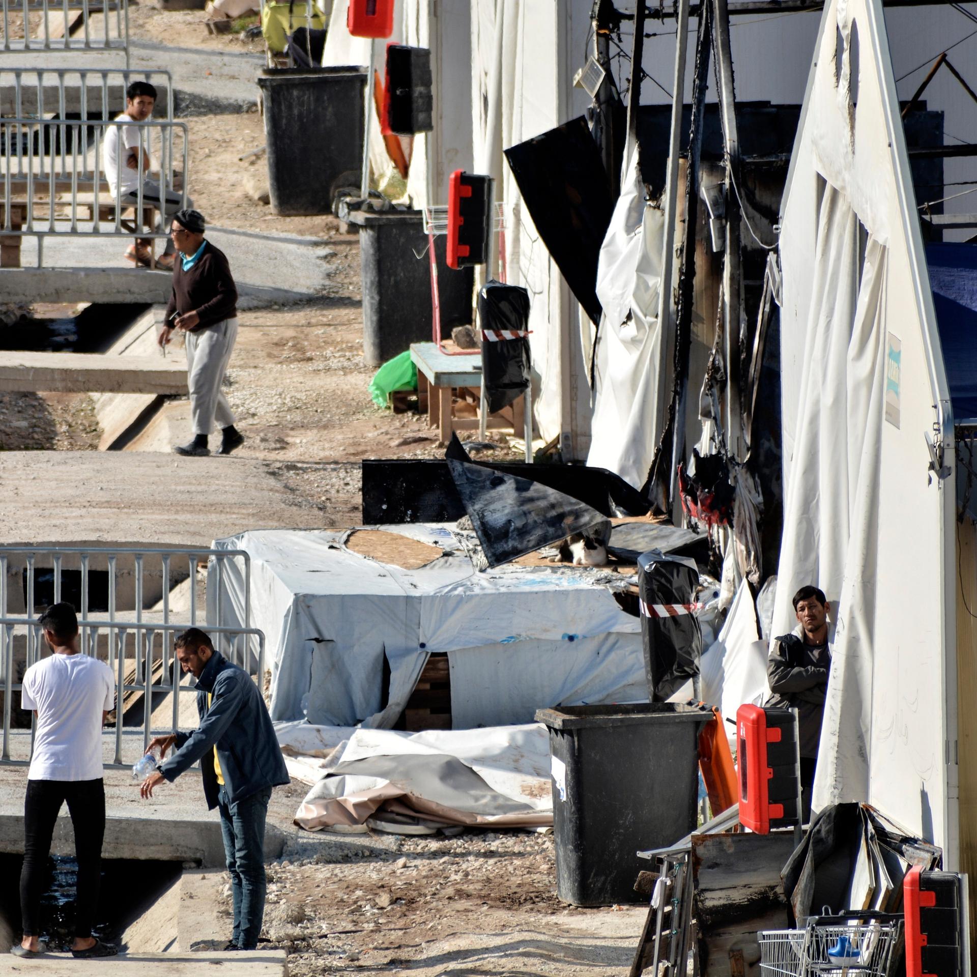 Flüchtlinge in einem Lager auf der Insel Lesbos stehen oder sitzen vor ihren Unterkünften.