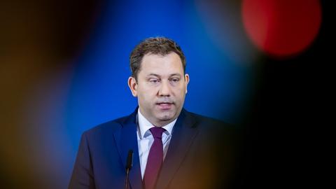 Klingbeil im schwarzen Jackett mit dunkler Krawatte. Er steht vor der blauen Wand der Bundespressekonferenz und sagt etwas. Im Vordergrund rechts und linkls am Bildrand unscharfe dunkle Silhouetten vermutlich von Zuhörern.