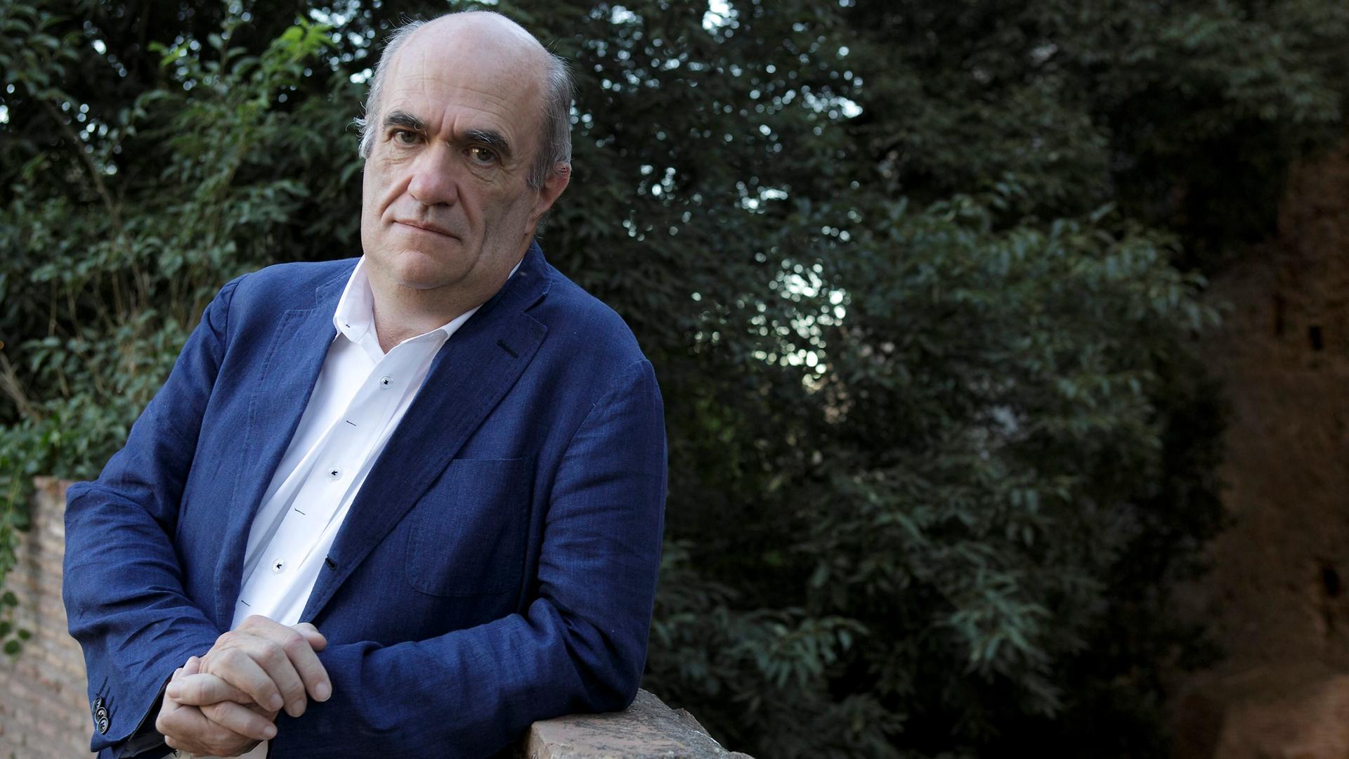 Der irische Schriftsteller Colm Tóibín steht an eine Mauer gelehnt. Hinter ihm sind Bäume.