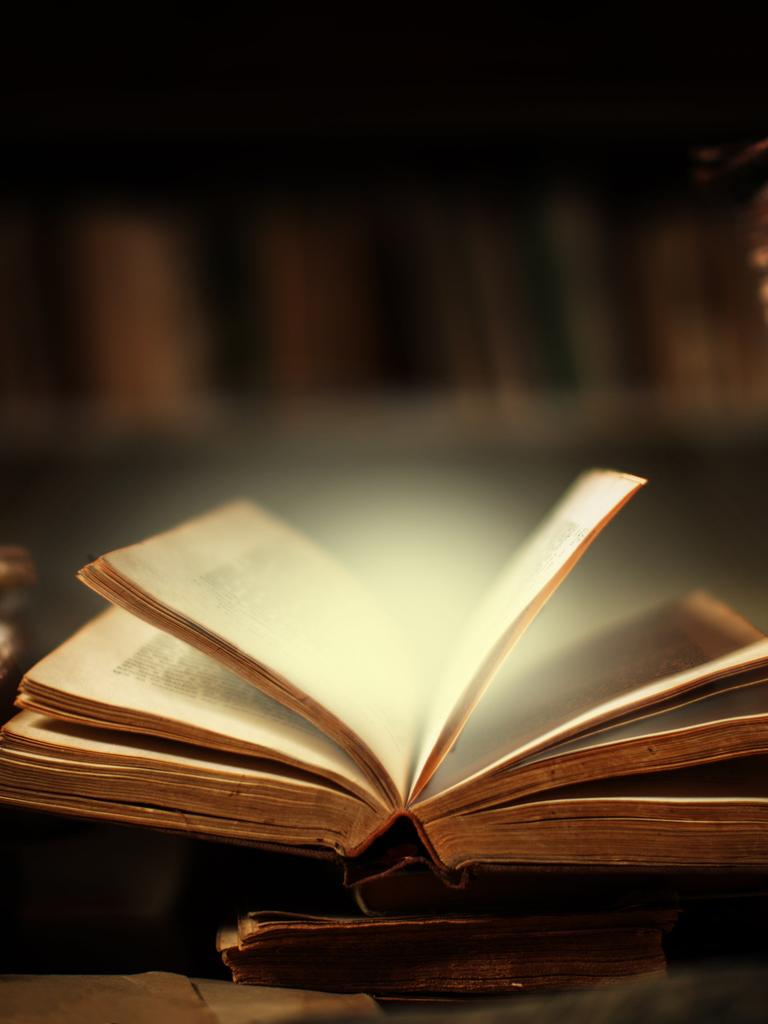 EIn Buch liegt offen auf dem Schreibtisch in der Bibliothek, die Seiten scheinen sich zu bewegen und erhellen die Umgebung durch ein magisches Leuchten