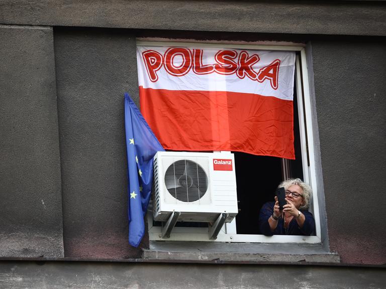 Eine Frau lehnt sich aus einem Fenster und fotografiert mit ihrem Handy eine Massendemostration der polnischen Opposition - über ihr hängt die polnische, neben ihr die europäische Flagge.