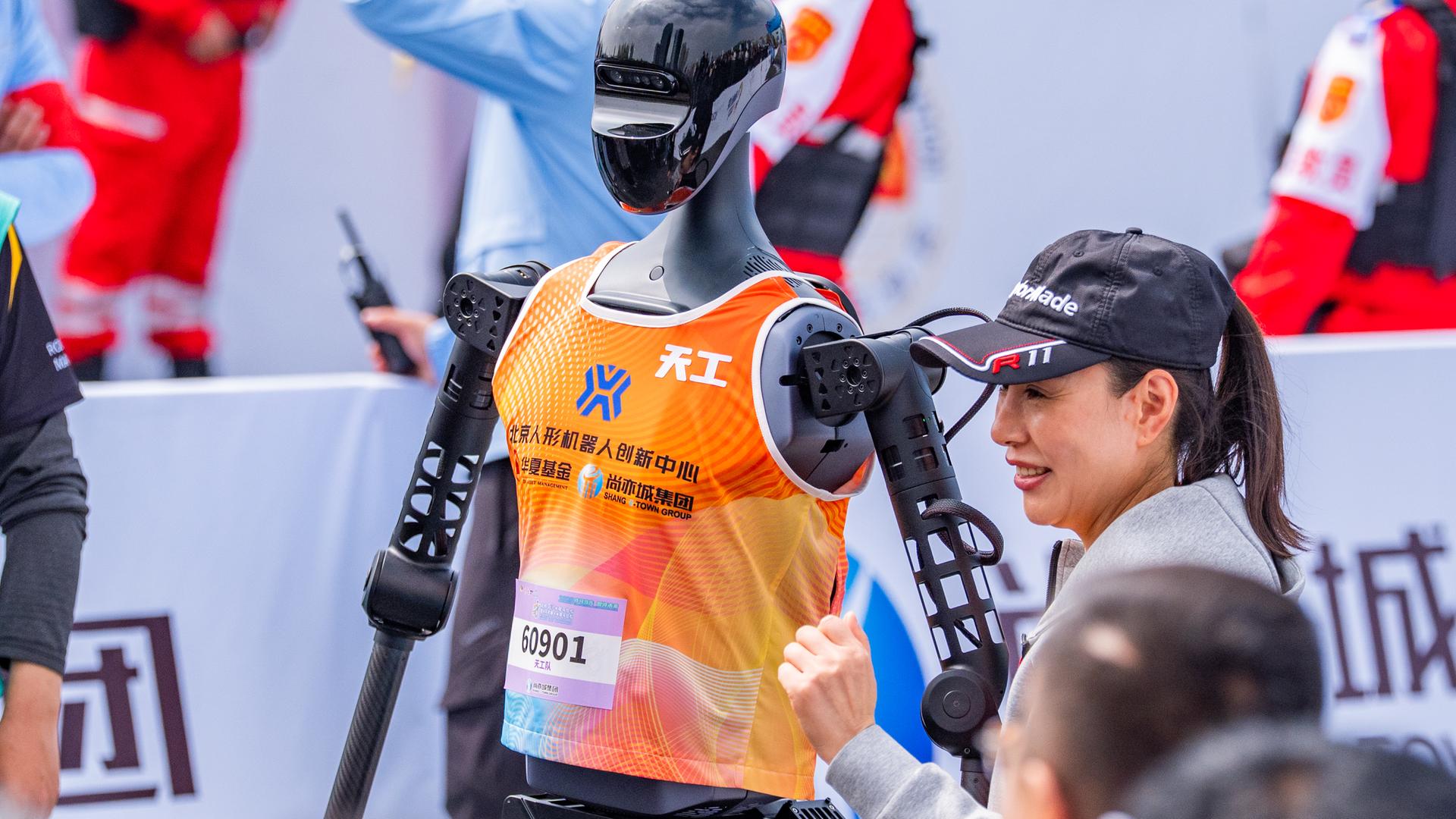 Eine Frau steht neben einem humanoiden Roboter mit orange farbenem Trägershirt, an welchem eine Startnummer befestigt ist. Eine Frau steht neben einem humanoiden Roboter mit orange farbenem Trägershirt, an welchem eine Startnummer befestigt ist.