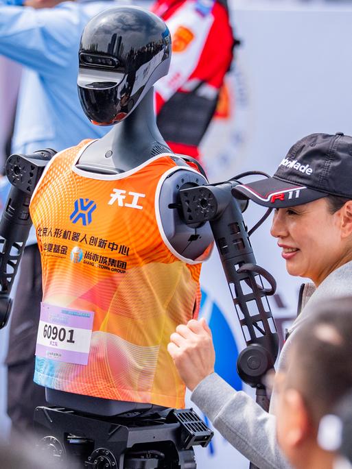 Eine Frau steht neben einem humanoiden Roboter mit orange farbenem Trägershirt, an welchem eine Startnummer befestigt ist.