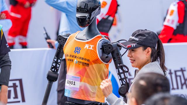 Eine Frau steht neben einem humanoiden Roboter mit orange farbenem Trägershirt, an welchem eine Startnummer befestigt ist.