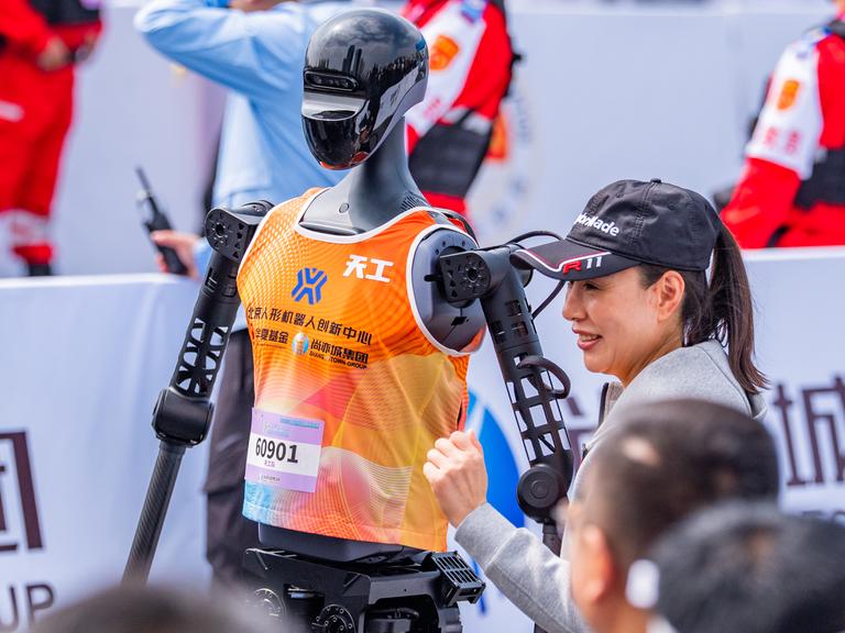 Eine Frau steht neben einem humanoiden Roboter mit orange farbenem Trägershirt, an welchem eine Startnummer befestigt ist.