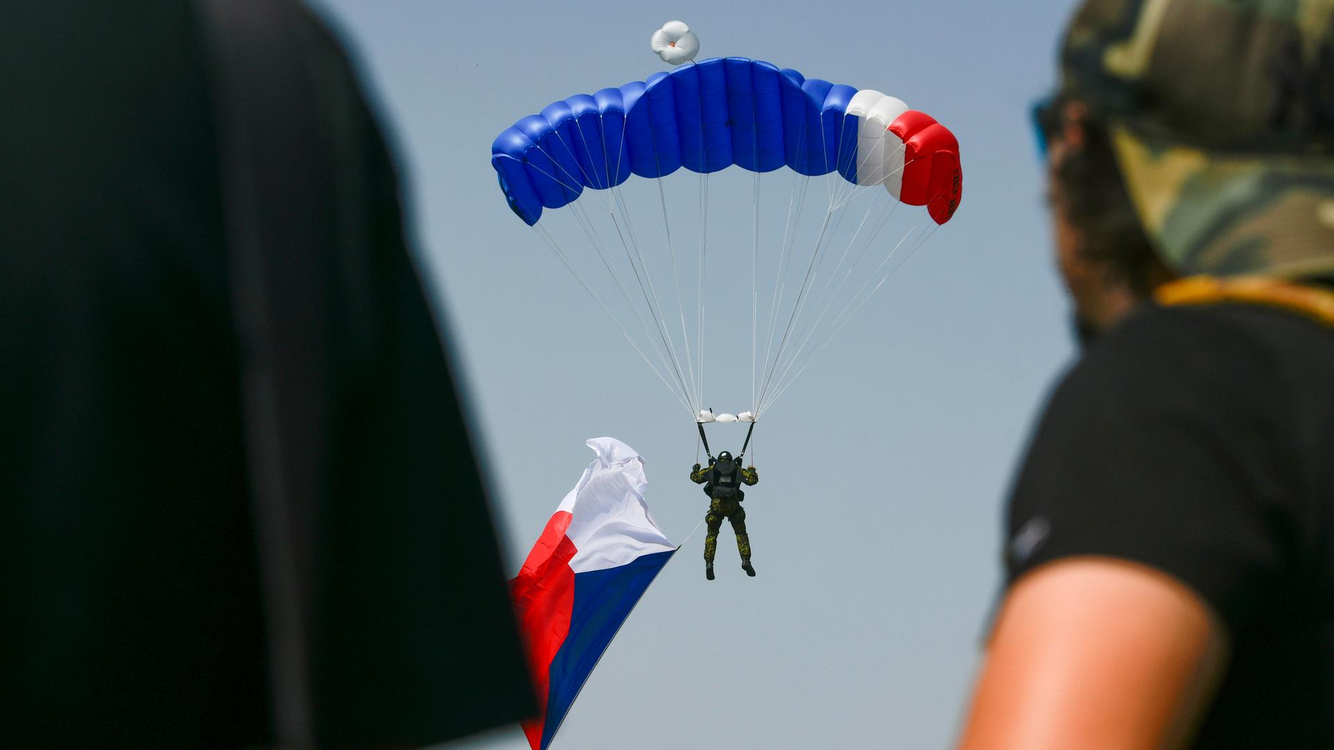 Ein Fallschimspringer mit einer tschechischen Flagge in der Hand landet zwischen Menschen, die dabei zuschauen. Der Fallschirm trägt ebenfalls die tschechischen Nationalfarben.