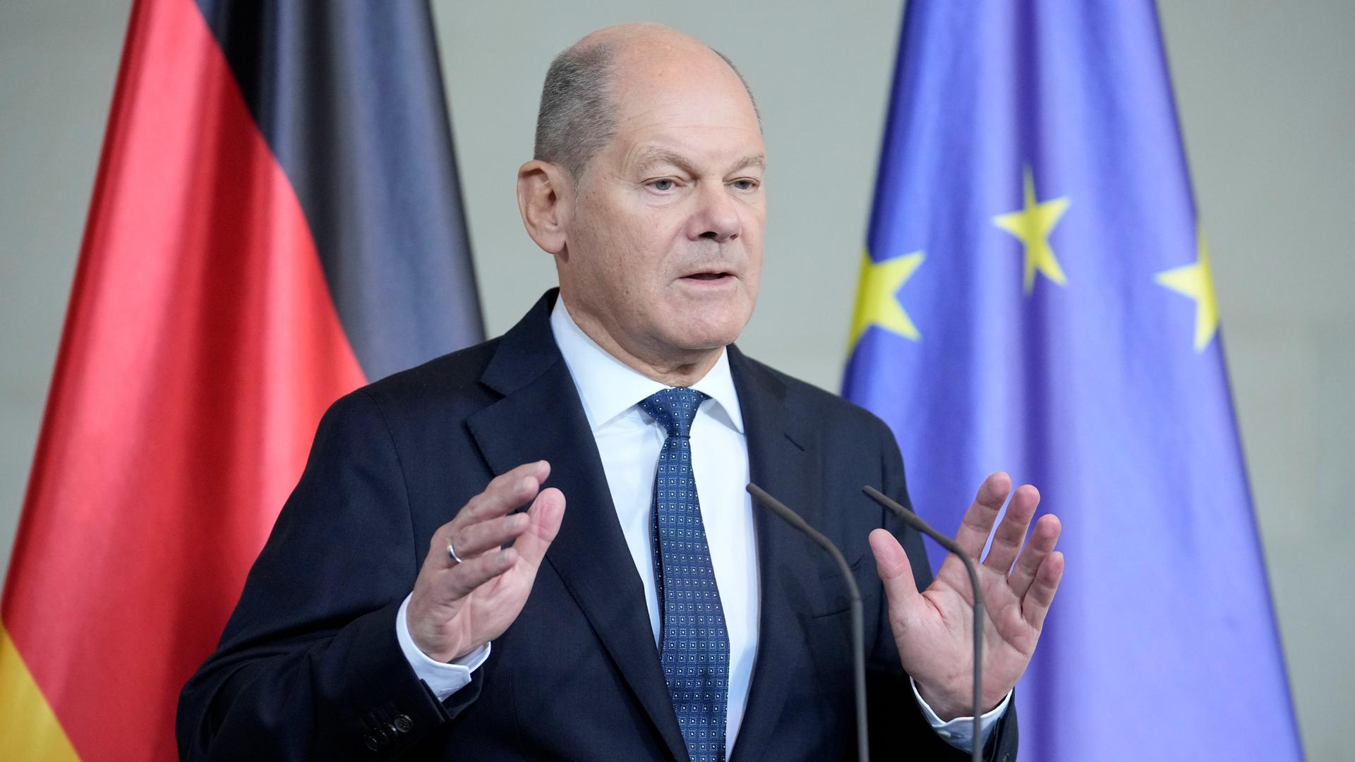 Olaf Scholz steht vor einer deutschen und einer EU-Flagge und spricht.