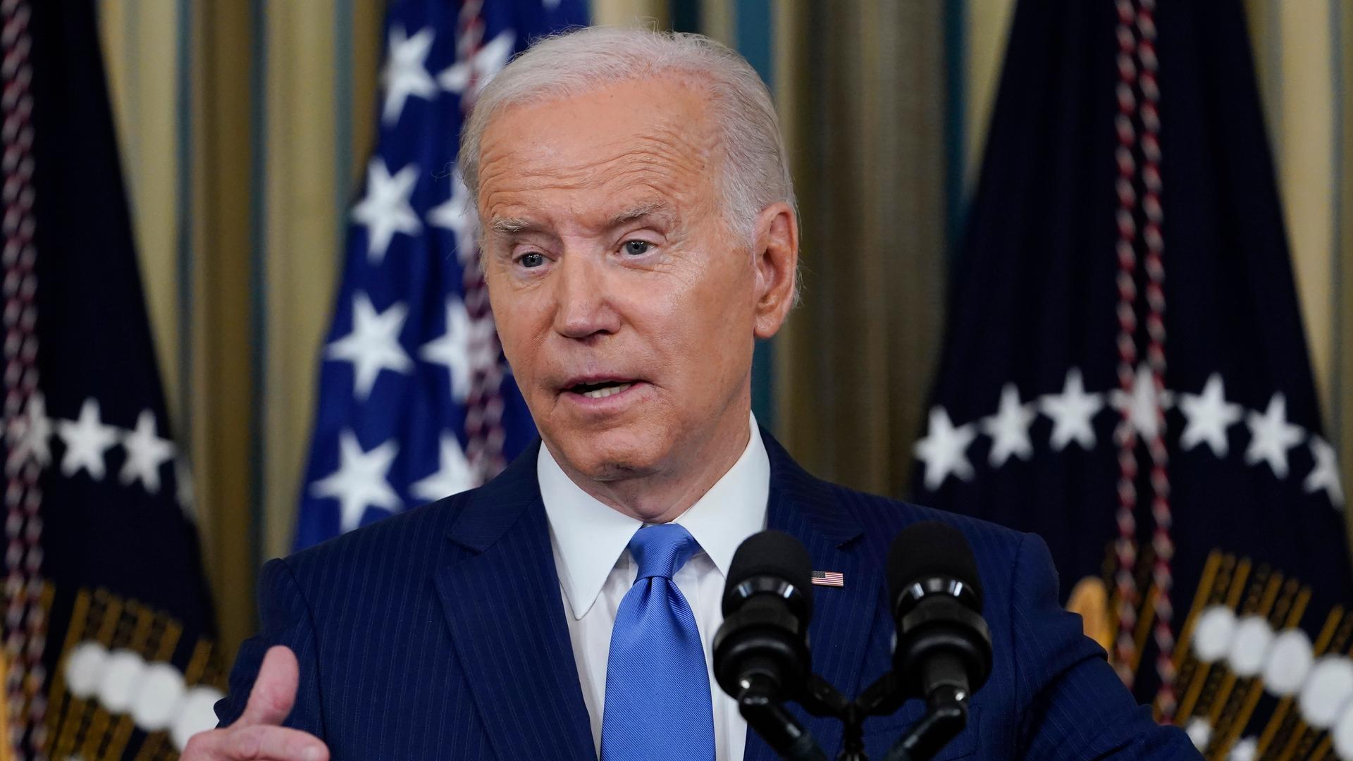Biden im dunkelblauen Anzug spricht in ein Mikrofon und gestikuliert mit der rechten Hand. Dahinter unscharf mehrere Flaggen. Biden im dunkelblauen Anzug spricht in ein Mikrofon und gestikuliert mit der rechten Hand. Dahinter unscharf mehrere Flaggen.