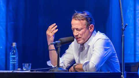 Ein Mann in weißem Hemd sitzt an einem Tisch mit Mikrofon. Er spricht mit geschlossenen Augen und gestikuliert mit einer Hand. Der Saal ist blau beleuchtet.