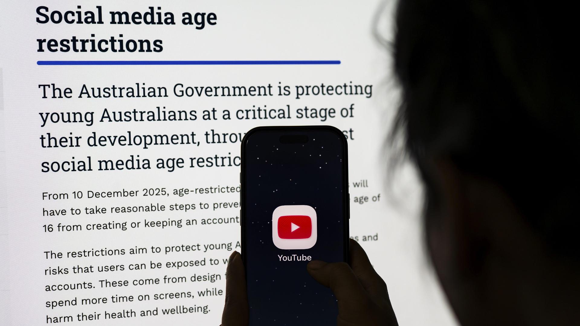 Eine Person hält Telefon mit der YouTube-App vor einen Bildschirm mit Informationen zu Australiens neuen Altersbeschränkungen für soziale Medien.