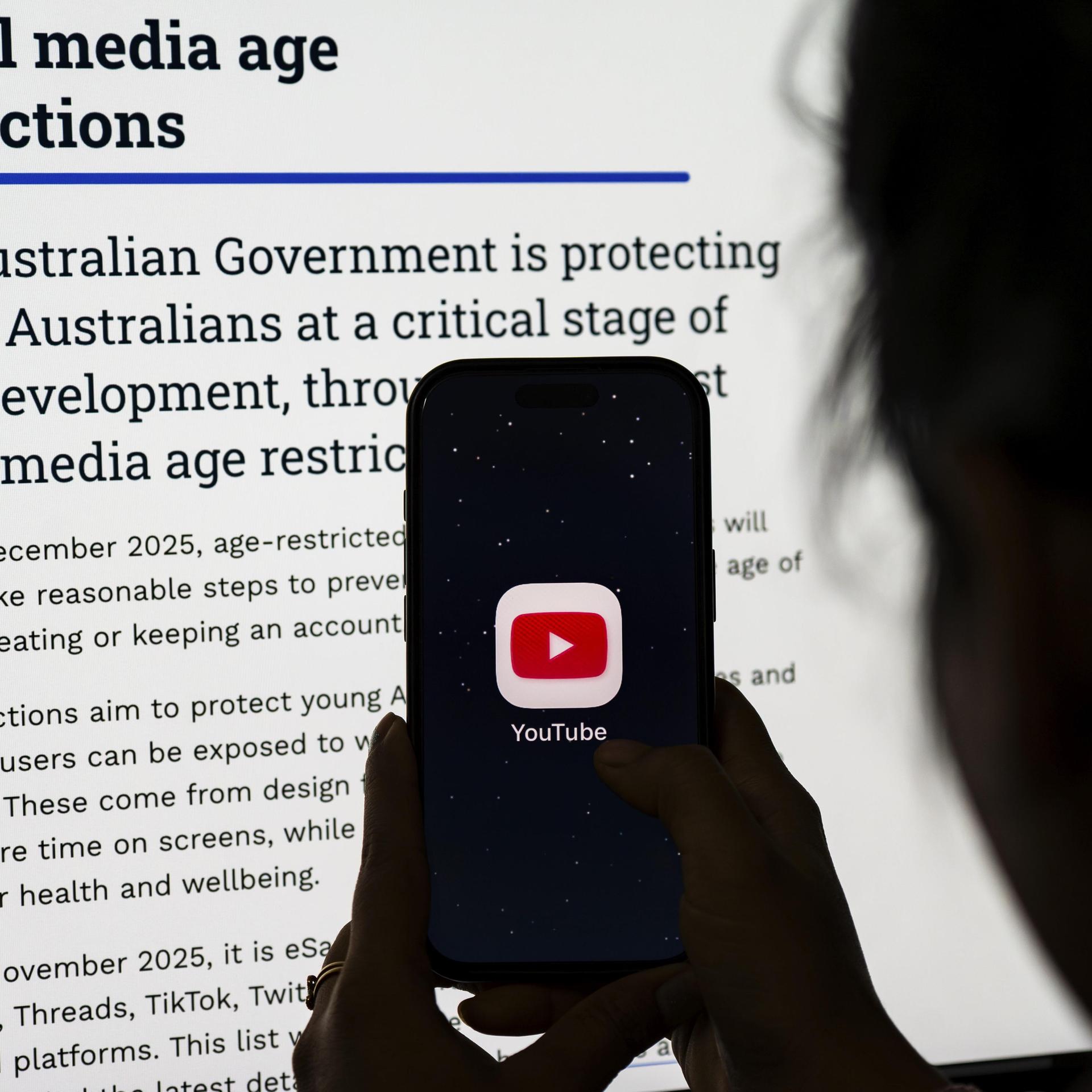 Eine Person hält Telefon mit der YouTube-App vor einen Bildschirm mit Informationen zu Australiens neuen Altersbeschränkungen für soziale Medien.
