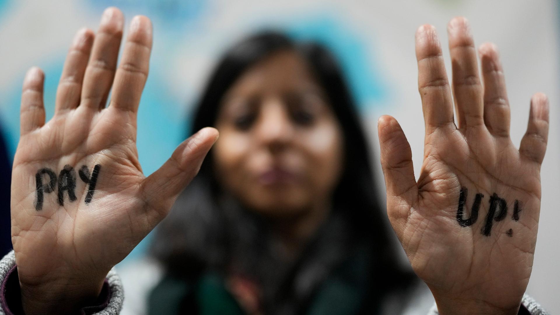 Die Frau hält beide Handflächen der Kamera entgegen. Ihr Gesicht ist nur verschwommen zu sehen. Auf den Handflächen steht "Pay up!", also "Zahlt!".