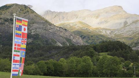 Blick auf die französischen Alpen bei Le Vernet im Sonnenschein, davor eine Fahne mit verschiedenen Flaggen, die an die Opfer des Pilotensuizids vor zehn Jahren erinnern sollen.