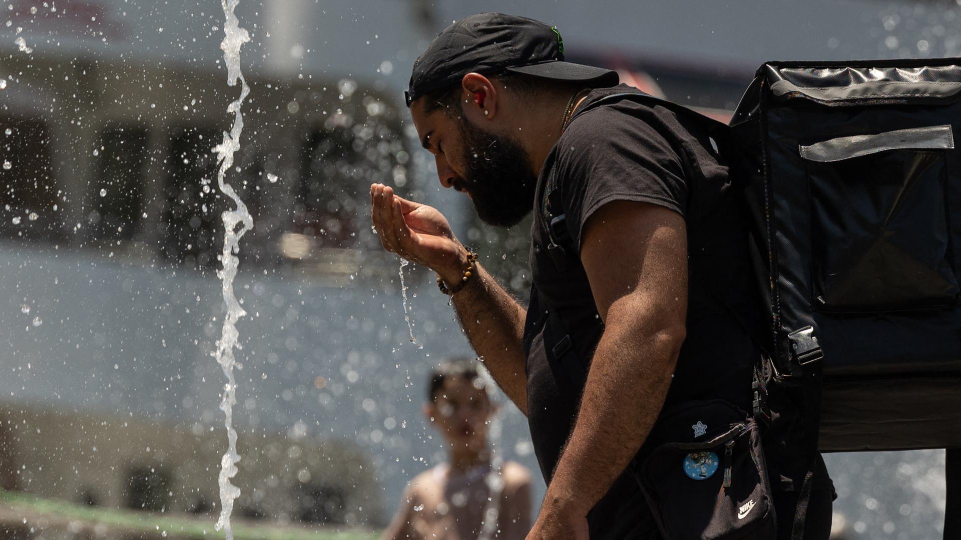 Ein bärtiger Mann in schwarzem T-Shirt und schwarzem Basecap mit einem großen schwarzen Rucksack trinkt Wasser aus einem Springbrunnen