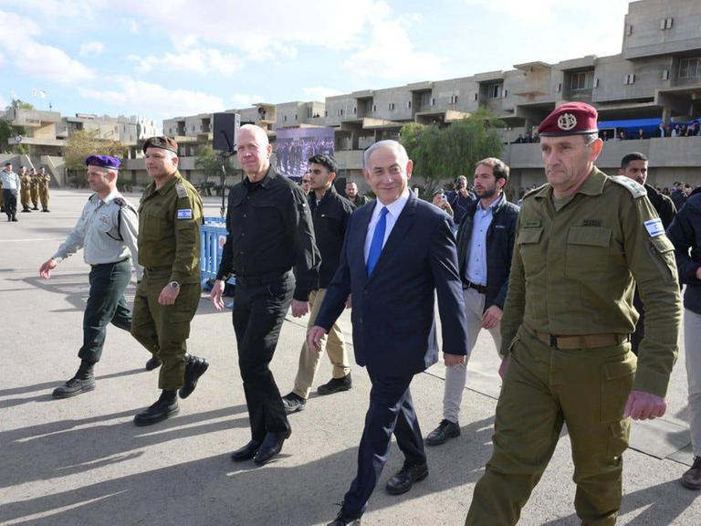 Zvilisten, Personenschützer und Männer in Uniformen schreiten durch den Staub. Die Uniformierten tragen rote Käppchen. Im Hintergrund ist eine Gruppe von Soldaten beim Exerzieren zu sehen.