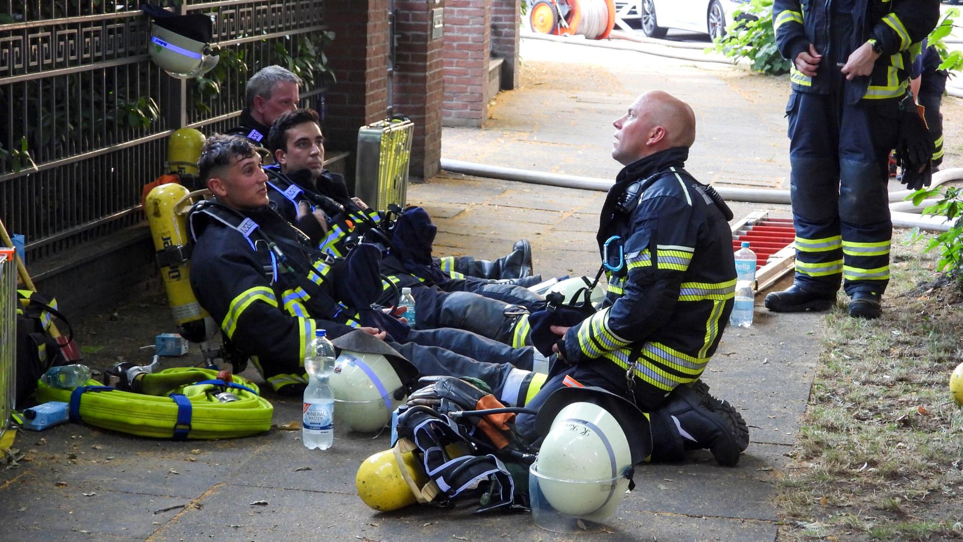 Vor Hitze erschöpfte Feuerwehrleute sitzen umgeben von Einsatzequipment auf einem Bürgersteig am Boden Vor Hitze erschöpfte Feuerwehrleute sitzen umgeben von Einsatzequipment auf einem Bürgersteig am Boden