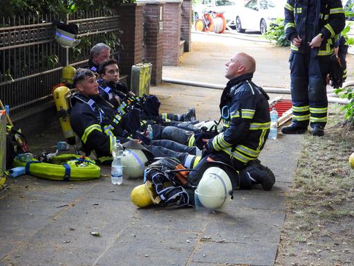Vor Hitze erschöpfte Feuerwehrleute sitzen umgeben von Einsatzequipment auf einem Bürgersteig am Boden