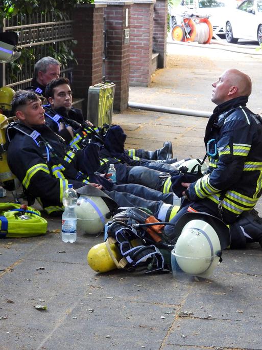 Vor Hitze erschöpfte Feuerwehrleute sitzen umgeben von Einsatzequipment auf einem Bürgersteig am Boden