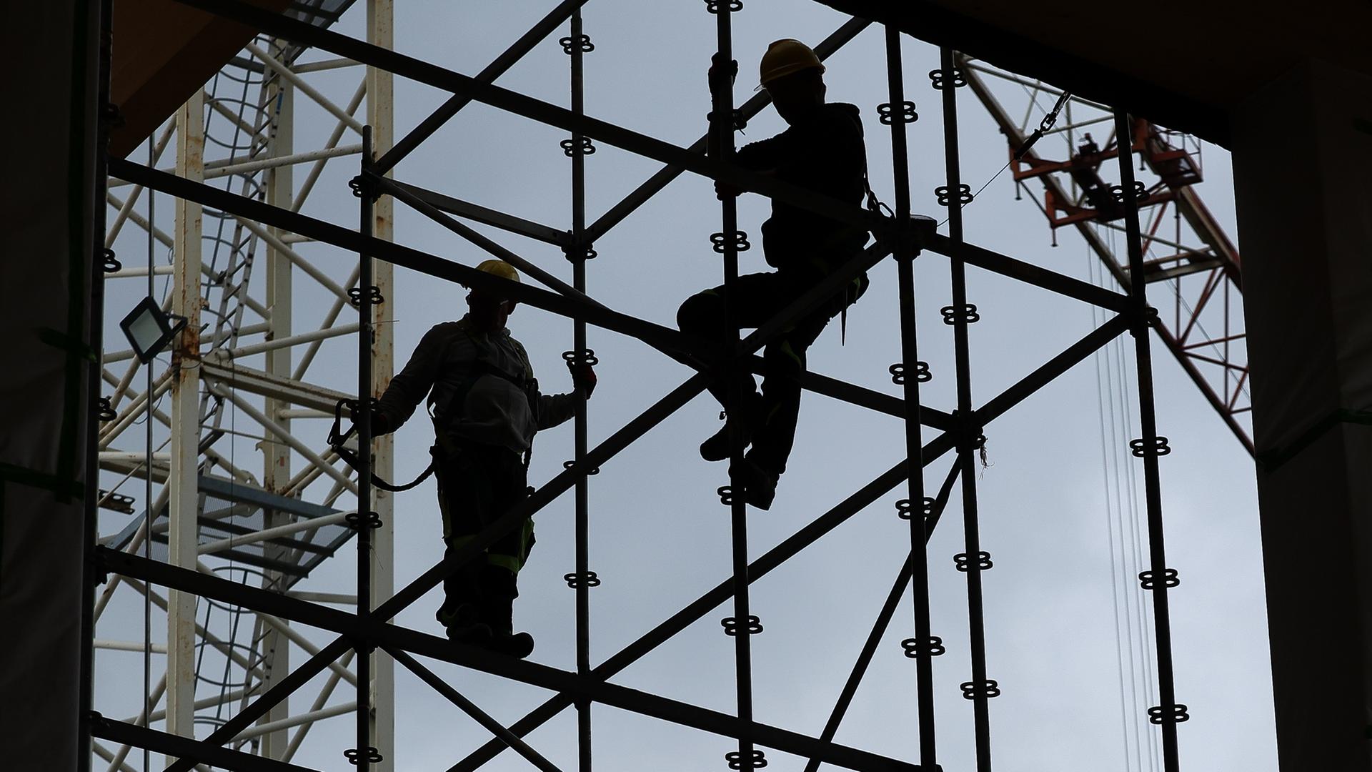 Gerüstbauer errichten mit Gurten gesichert ein Gerüst auf der Baustelle. Hinter ihnen der blaue Himmel, das Gerüst und die Personen sind nur als Silhouetten sichtbar.