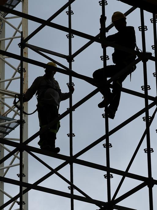 Gerüstbauer errichten mit Gurten gesichert ein Gerüst auf der Baustelle. Hinter ihnen der blaue Himmel, das Gerüst und die Personen sind nur als Silhouetten sichtbar.