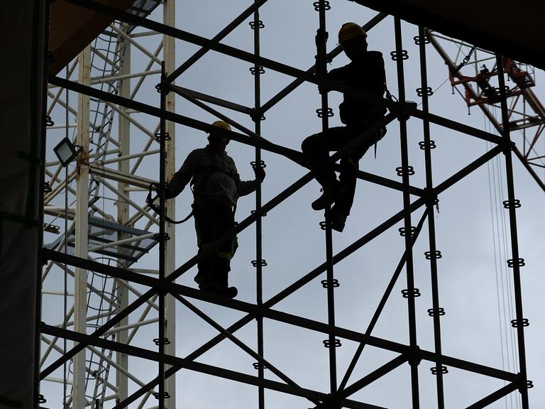 Gerüstbauer errichten mit Gurten gesichert ein Gerüst auf der Baustelle. Hinter ihnen der blaue Himmel, das Gerüst und die Personen sind nur als Silhouetten sichtbar.