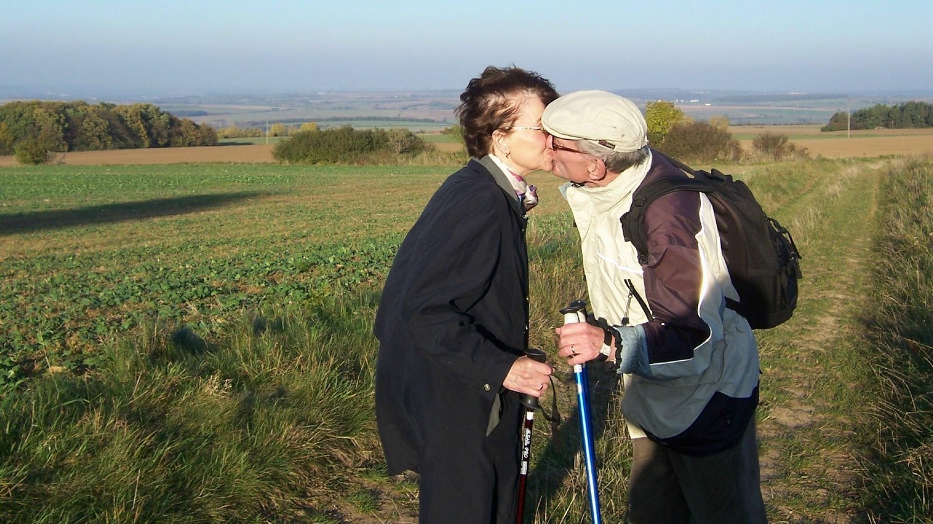 Ein älteres Paar steht mit Nordic-Walking-Stöcken küssend auf einem Feld.