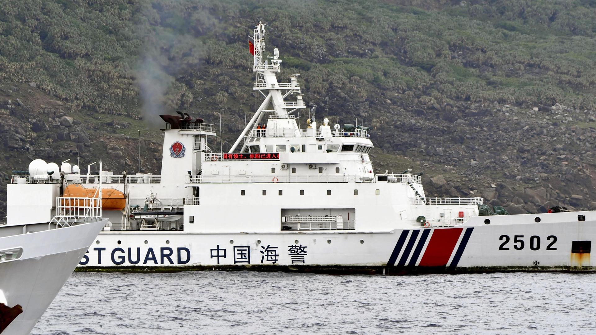 Das weiße Schiff mit der Aufschrift "Cost Guard" liegt vor der Küste, die man im Hintergrund als einen mit Büschen bewachsenen steinigen Hang sieht.