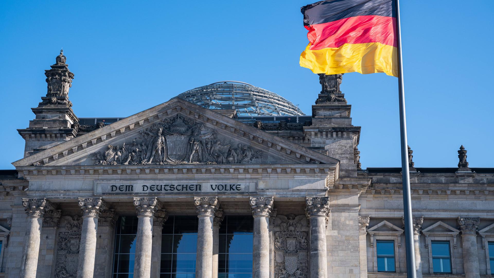 Westfassade des Reichstagsgebäudes mit dem Schriftzug Dem Deutschen Volke und einer Nationalflagge davor.
