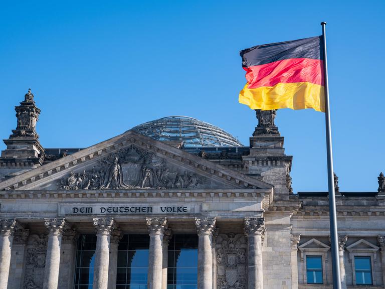 Westfassade des Reichstagsgebäudes mit dem Schriftzug Dem Deutschen Volke und einer Nationalflagge davor.