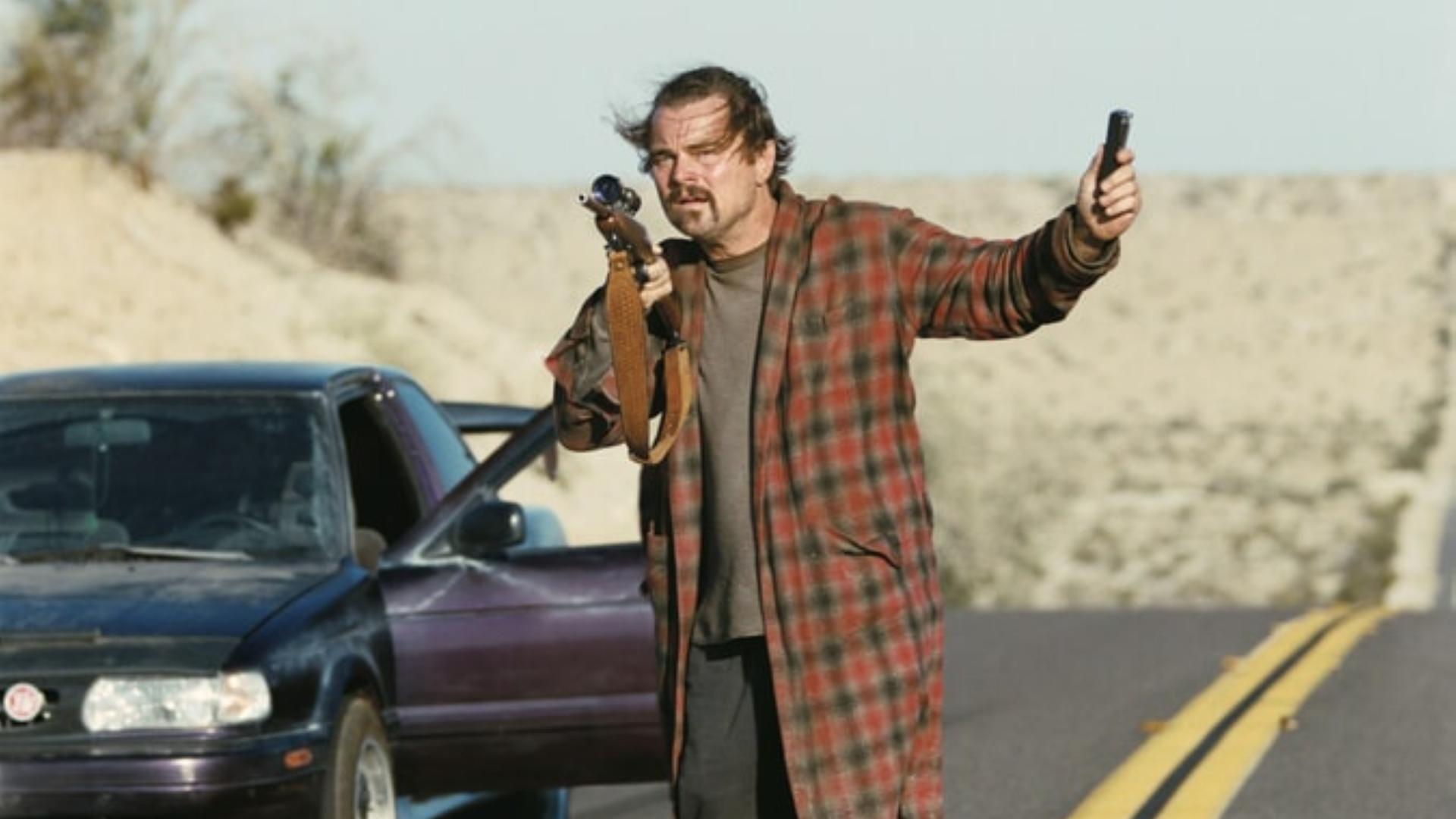Leonardo DiCaprio hält in einer Filmszene eine Pistole in die Luft während er neben einem Auto steht.