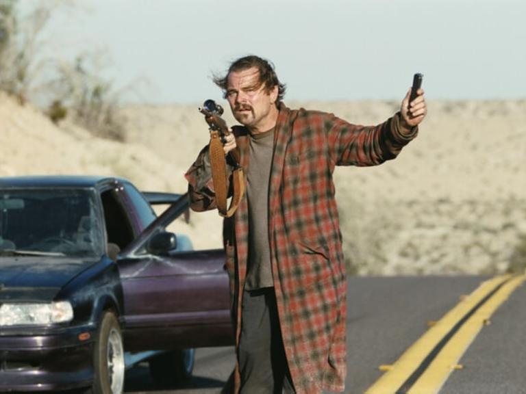 Leonardo DiCaprio hält in einer Filmszene eine Pistole in die Luft während er neben einem Auto steht.