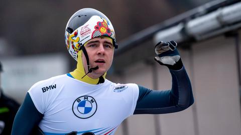 Der ukrainische Skeleton-Pilot Vladyslav Heraskevych steht auf der Skeleton-Bahn in Winterberg. Er hat einen Anzug und seinen Helm auf, aber sein Gesicht ist zu sehen. Seine linke Hand ist erhoben. 