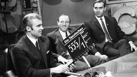 Auf einem Schwarz-Weiß-Foto sitzt Uwe Seeler zwischen zwei Moderatoren im Studio der Sportschau. Der Moderator links hält eine Film-Klappe ins Bild.