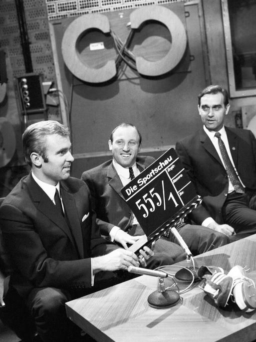 Auf einem Schwarz-Weiß-Foto sitzt Uwe Seeler zwischen zwei Moderatoren im Studio der Sportschau. Der Moderator links hält eine Film-Klappe ins Bild.