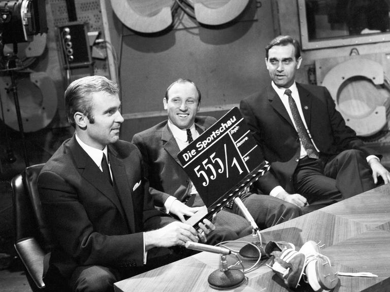 Auf einem Schwarz-Weiß-Foto sitzt Uwe Seeler zwischen zwei Moderatoren im Studio der Sportschau. Der Moderator links hält eine Film-Klappe ins Bild.