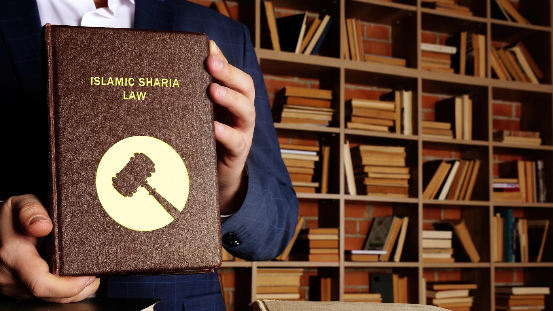 Ein Mann hält mit zwei Händen die Scharia, das islamische Recht fest, im Hintergrund in Regalen sieht man noch viele Bücher.