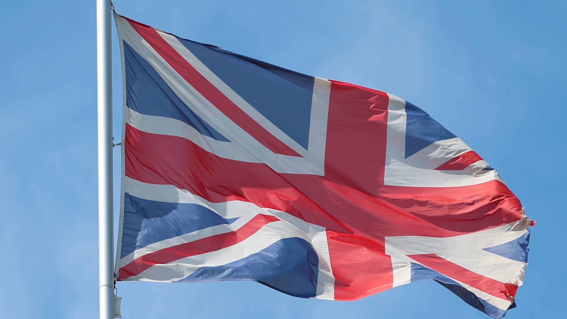Die britische Fahne von Großbritannien weht an einem Fahnenmast vor blauem Himmel im Wind. Die britische Fahne von Großbritannien weht an einem Fahnenmast vor blauem Himmel im Wind.