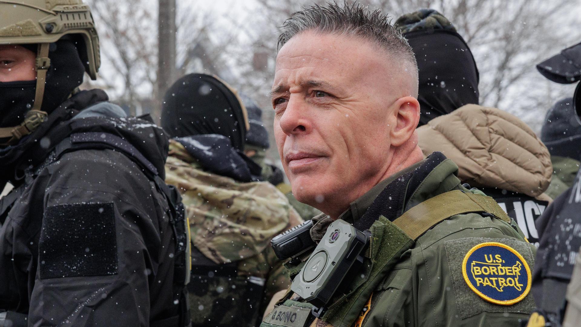 ICE-Einsatzleiter Gregory Bovino steht in Uniform vor zwei vermummten ICE-Männern