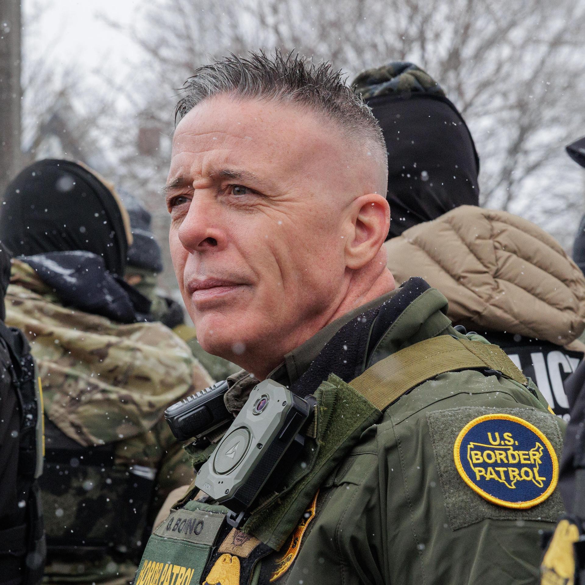 ICE-Einsatzleiter Gregory Bovino steht in Uniform vor zwei vermummten ICE-Männern