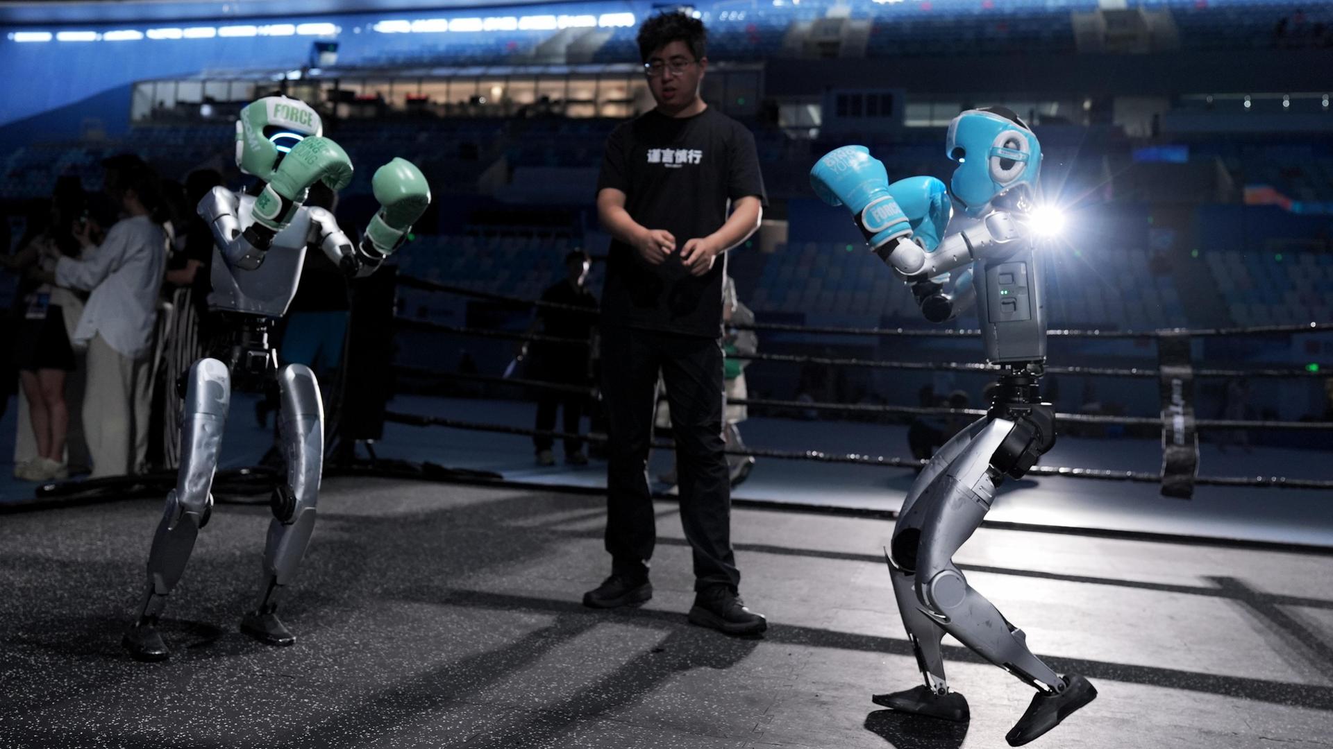Ein Mensch steht zwischen zwei Robotern mit Boxhandschuhen, die gegeneinander kämpfen sollen. 