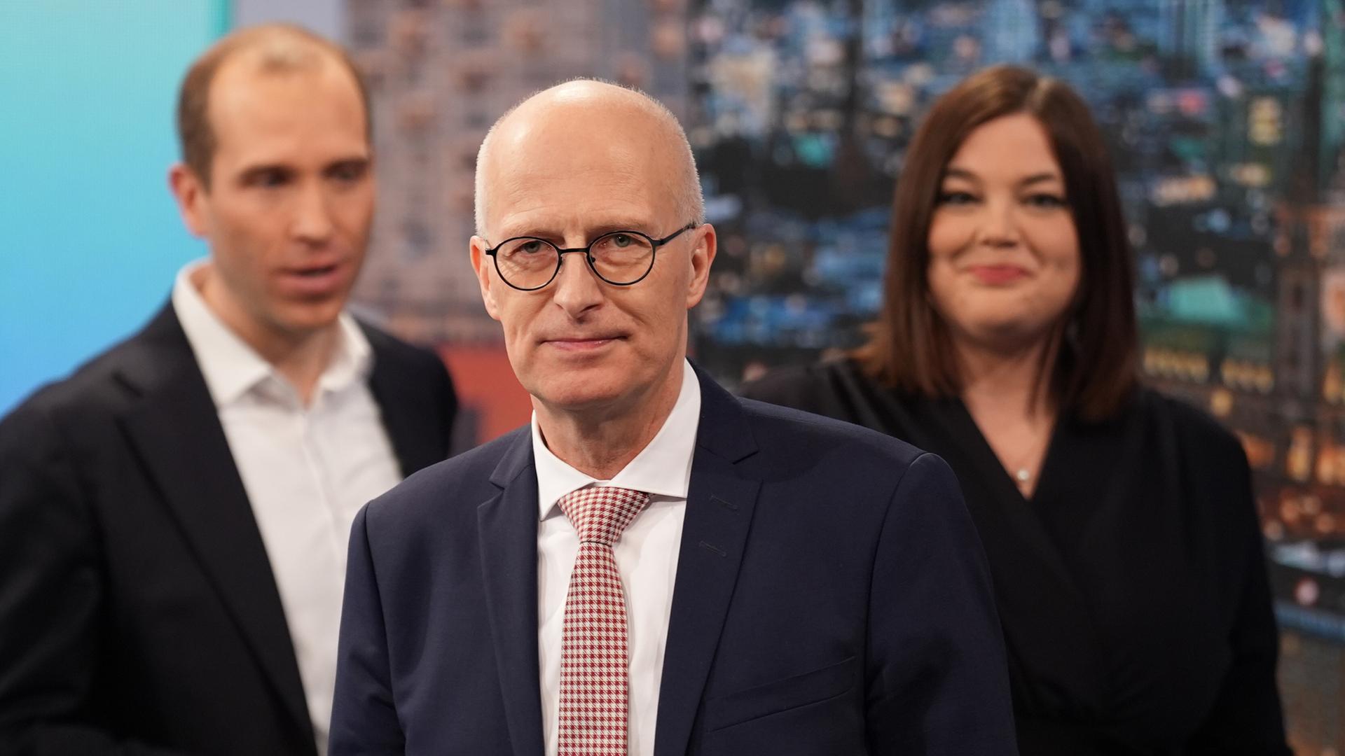 Peter Tschentscher steht lächelnd in einem Fernsehstudio. Hinter ihm sind leicht unscharf Dennis Thering und Katharina Fegebank zu sehen.