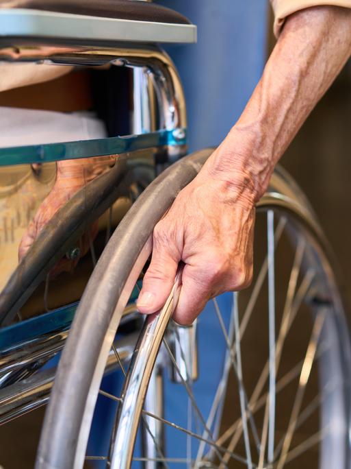 Nahaufnahme: Ältere Person hält Rollstuhlrad im Korridor einer Rehabilitationseinrichtung Nahaufnahme: Ältere Person hält Rollstuhlrad im Korridor einer Rehabilitationseinrichtung