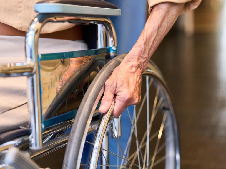 Nahaufnahme: Ältere Person hält Rollstuhlrad im Korridor einer Rehabilitationseinrichtung Nahaufnahme: Ältere Person hält Rollstuhlrad im Korridor einer Rehabilitationseinrichtung