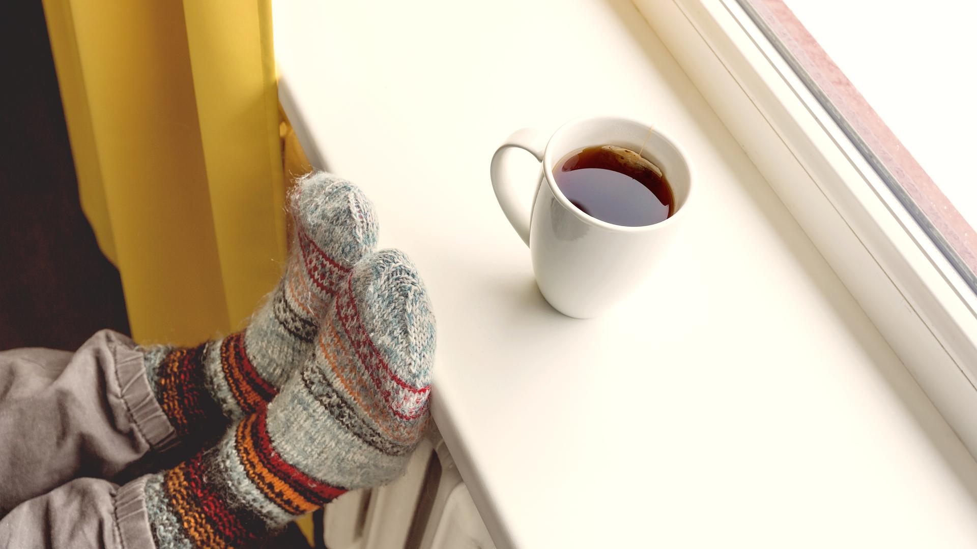 Eine Tasse mit heissem Tee steht auf einer Fensterbank, Füße in Wollsocken liegen auf der Heizung.