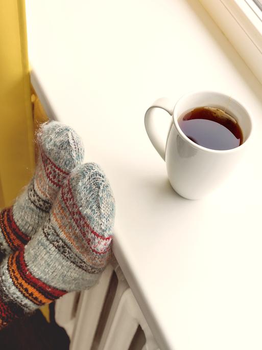 Eine Tasse mit heissem Tee steht auf einer Fensterbank, Füße in Wollsocken liegen auf der Heizung.