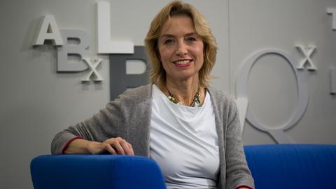 Susanne Kippenberger sitzt auf einem blauen Sofa und lächelt in die Kamera. Sie hat schulterlanges blondes Haar und trägt eine graue Strickjacke. Susanne Kippenberger sitzt auf einem blauen Sofa und lächelt in die Kamera. Sie hat schulterlanges blondes Haar und trägt eine graue Strickjacke.