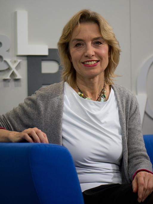 Susanne Kippenberger sitzt auf einem blauen Sofa und lächelt in die Kamera. Sie hat schulterlanges blondes Haar und trägt eine graue Strickjacke.