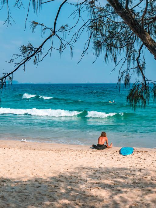 Ein Surfer sitzt mit seinem Surfbrett am Rainbow Bay Beach in Australien.